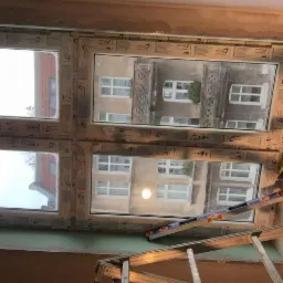 Montaż okna PCV w mieszkaniu, widoczny poziomica i drabina, w tle budynek z oknami i zielenią.