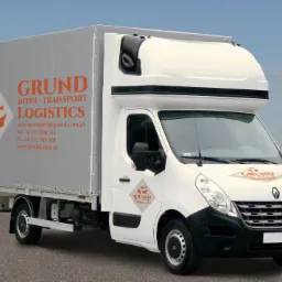 Biała furgonetka transportowa z szarą plandeką, na kt&oacute;rej widnieje pomarańczowe logo firmy Grund Inter-Transport Logistics wraz z danymi kontaktowymi, zaparkowana na asfaltowej drodze na tle pola...