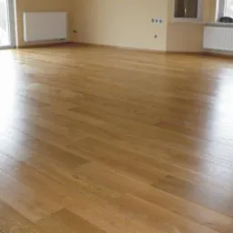 Jasna, parkietowa podłoga z wyraźnym połyskiem w pustym pokoju z beżowymi ścianami, białymi grzejnikami i oknami.