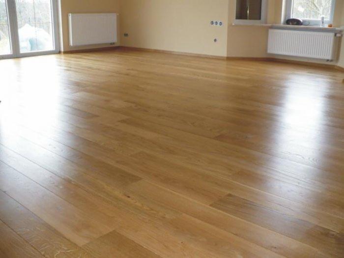 Jasna, parkietowa podłoga z wyraźnym połyskiem w pustym pokoju z beżowymi ścianami, białymi grzejnikami i oknami.