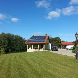 Dom jednorodzinny z panelami słonecznymi na dachu, widok z trawnika, samochód z otwartym bagażnikiem na podjeździe, błękitne niebo.