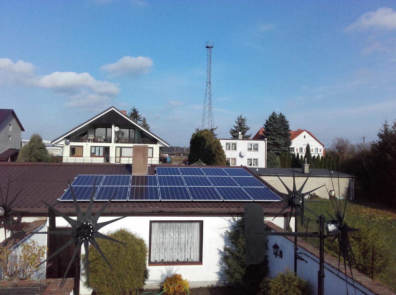 Instalacja paneli słonecznych na brązowym dachu domu jednorodzinnego, widok z góry, w tle inne budynki i wieża telekomunikacyjna.