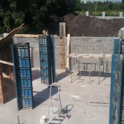 Budowa domu w trakcie realizacji: widoczne szalunki, ściany z cegły i bloczków betonowych, podłoże betonowe, drabina aluminiowa, w tle hałda ziemi i zaparkowane samochody.