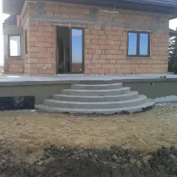 Surowy stan budynku z pustaków ceramicznych z widocznym fundamentem, izolacją przeciwwilgociową i betonowymi schodami wejściowymi, teren wokół budynku częściowo wyrównany.