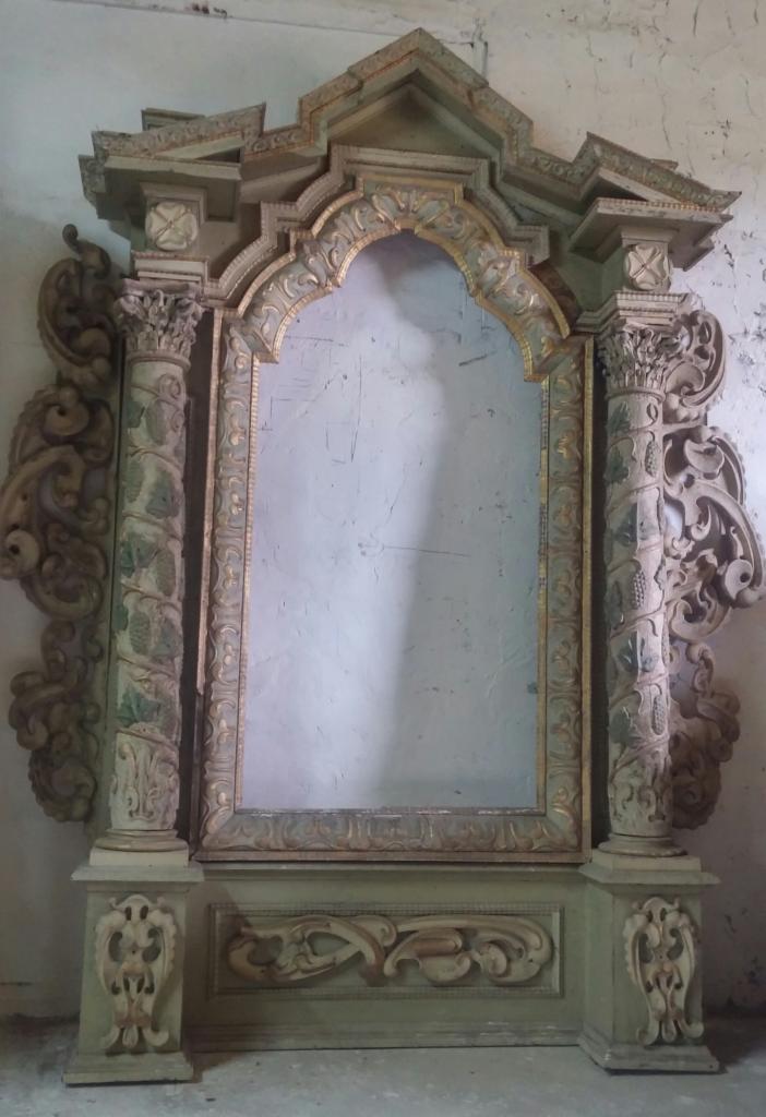 Antyczne, bogato zdobione lustro w drewnianej ramie z kolumnami, widoczne ślady renowacji, szara ściana w tle.