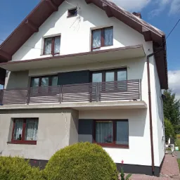 Dwupiętrowy dom jednorodzinny z białą elewacją i brązowym dachem, widoczny po zakończeniu prac, z elementami w kolorze grafitowym, brązowe okna i balustrady, na pierwszym planie zielone krzewy,...