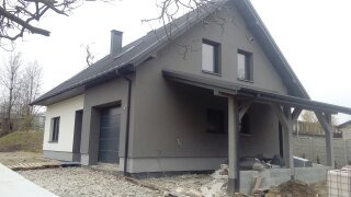 Nowoczesny dom jednorodzinny z szarą elewacją, widoczne elementy wykończeniowe, boczna wiata z drewnianą konstrukcją, teren wokół domu częściowo utwardzony żwirem.