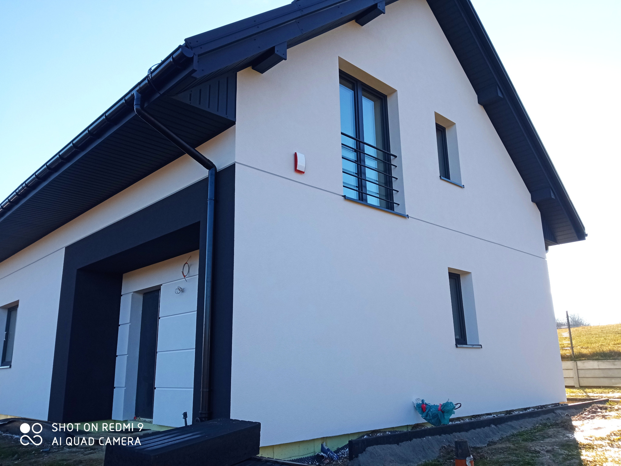 Nowoczesny dom jednorodzinny z jasną elewacją, czarnymi elementami wykończeniowymi dachu i okien, oraz widocznymi pozostałościami po pracach budowlanych wokół budynku.