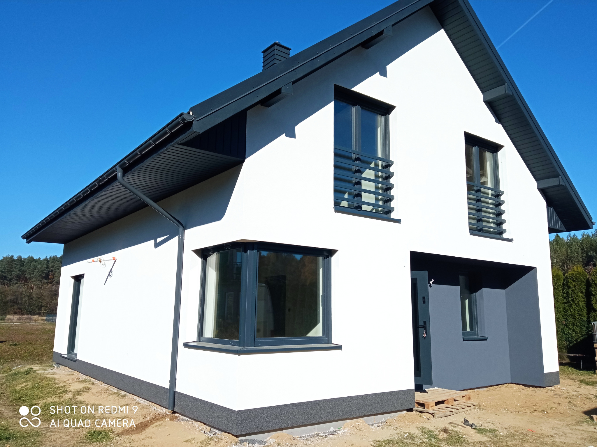 Nowoczesny dom jednorodzinny z białą elewacją, grafitowymi elementami wykończeniowymi i widocznymi śladami prac budowlanych wokół fundamentów.