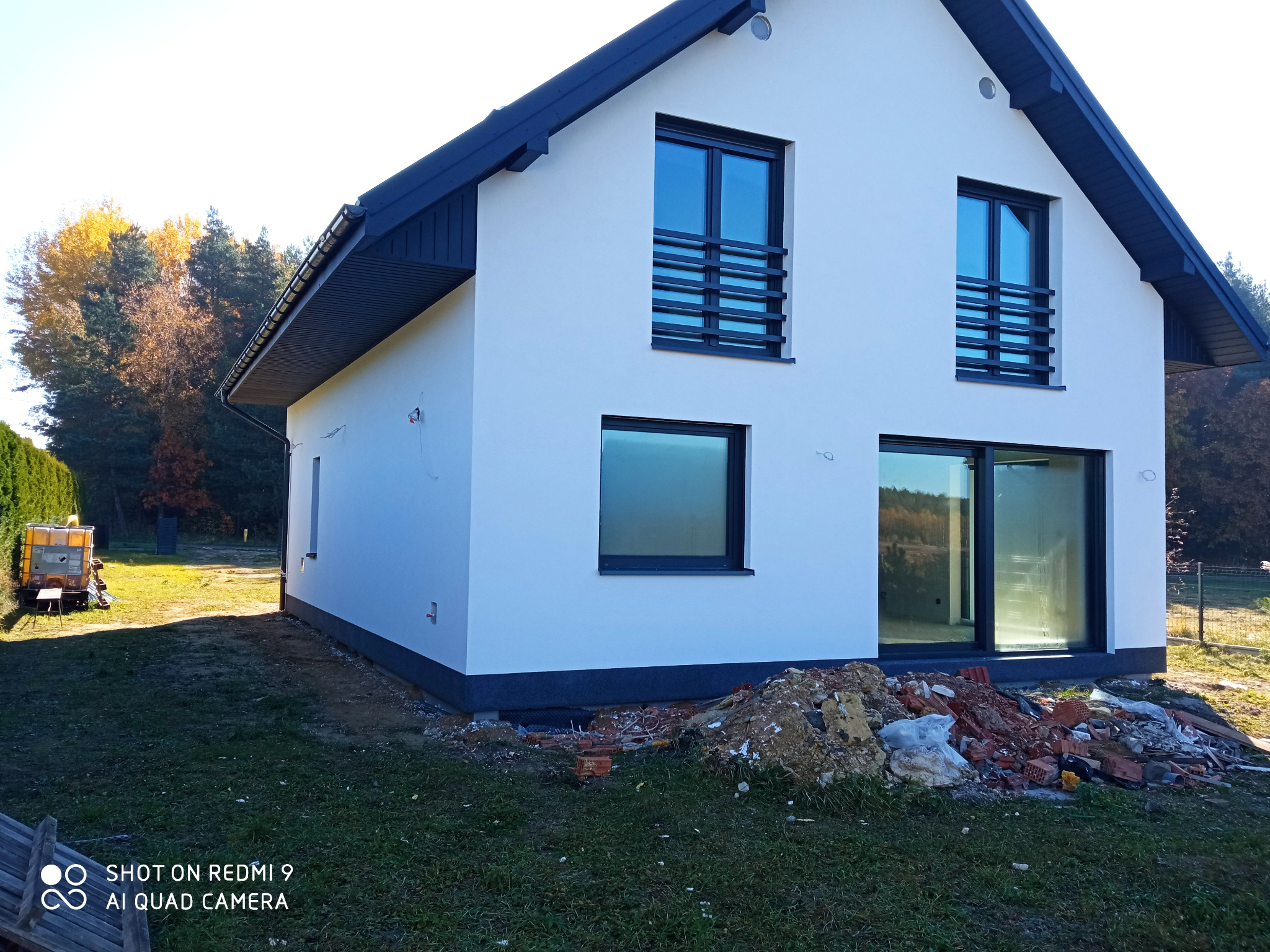 Nowoczesny, dwukondygnacyjny dom z białą elewacją i ciemnymi oknami oraz dachem, z widocznym stosem gruzu i odpadów budowlanych przy ścianie, sugerującym trwające prace wykończeniowe.