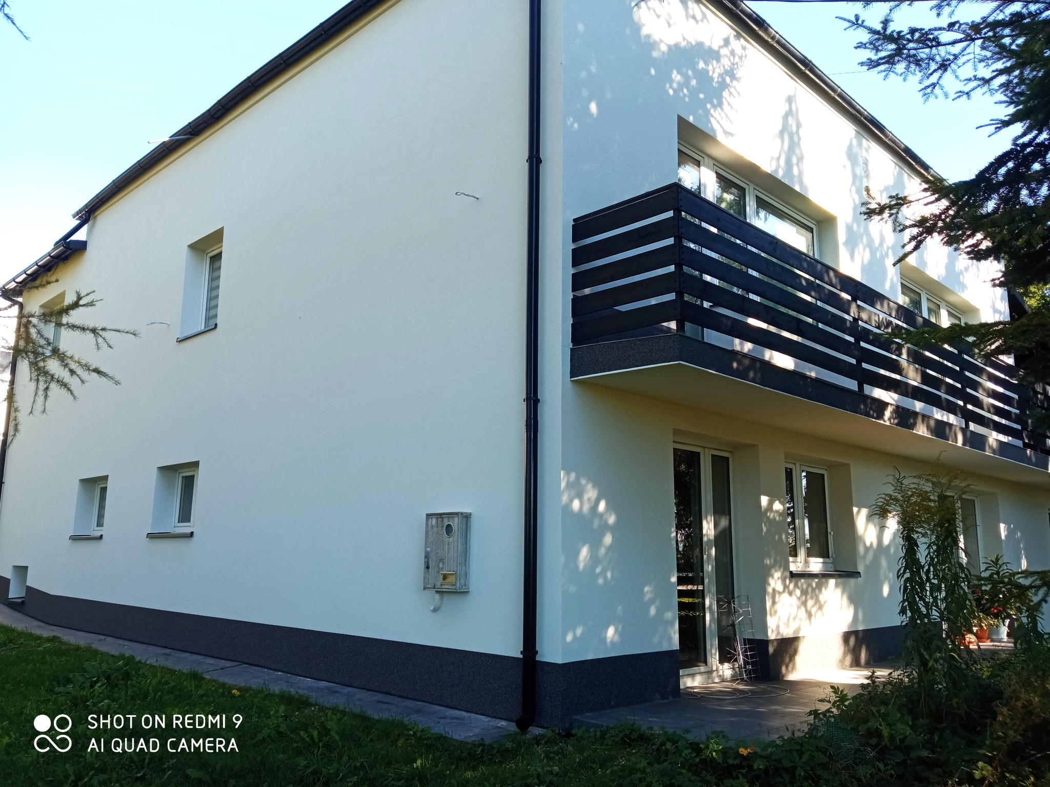 Dwukondygnacyjny dom z białą elewacją i ciemnym cokołem, balkon z czarną, poziomą balustradą, widoczne okna i drzwi balkonowe, na ścianie skrzynka na listy, w tle zieleń drzew i trawnik.