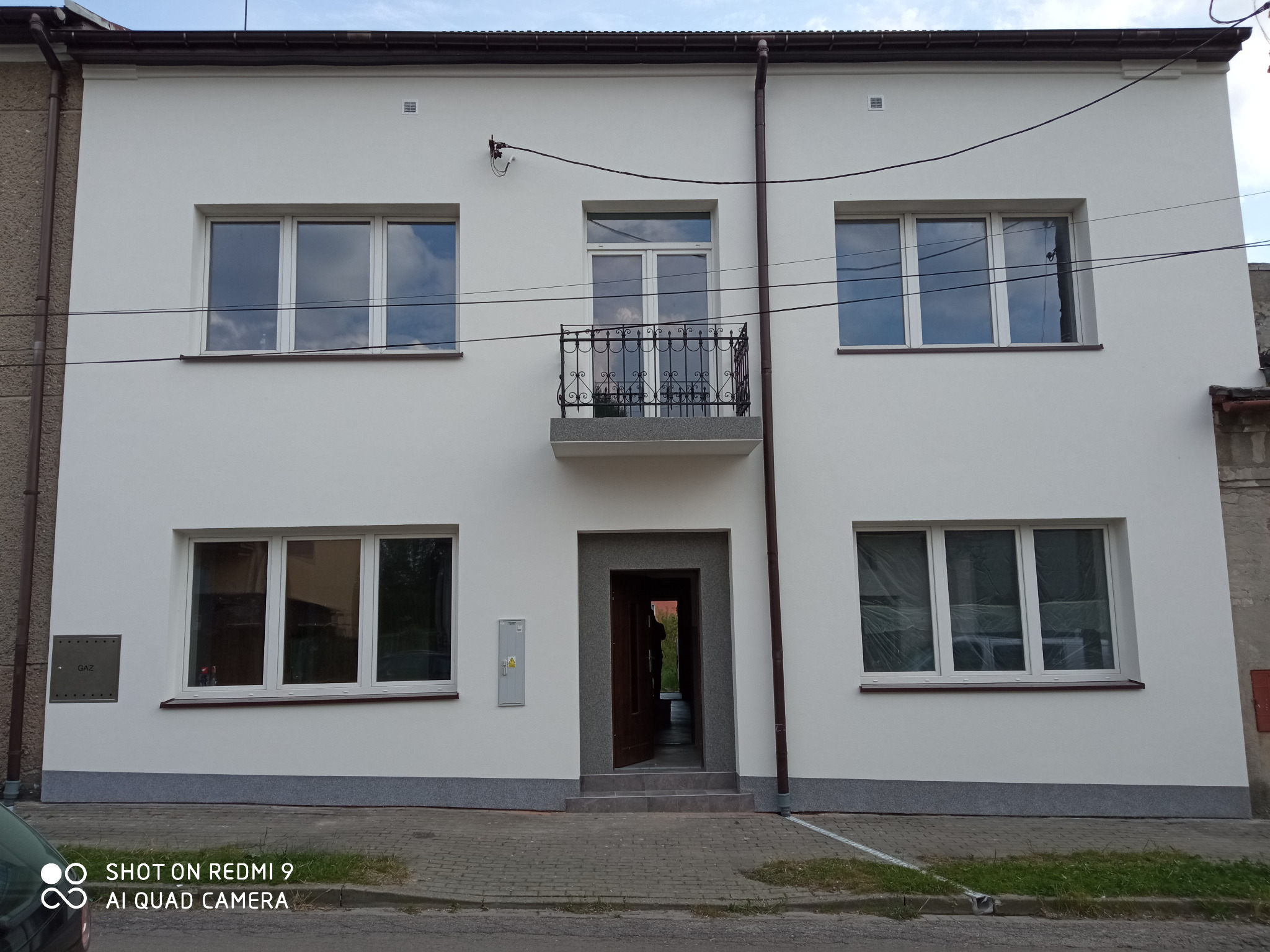 Odnowiony dwukondygnacyjny budynek z białą elewacją, szarym cokołem i balkonem z ozdobną, kutą balustradą. Widoczne okna z białymi ramami, brązowe rynny i skrzynka gazowa na ścianie.