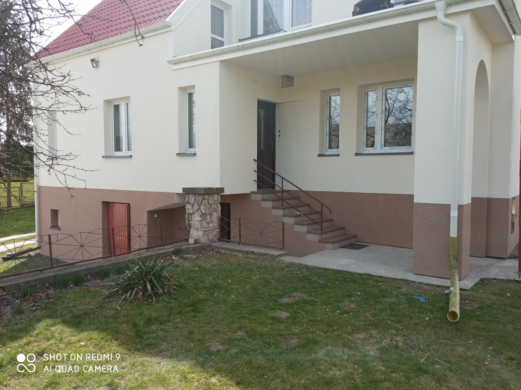 Dwukondygnacyjny dom z jasną elewacją, brązowym cokołem i czerwonym dachem, widoczny z poziomu trawnika z przodu budynku.