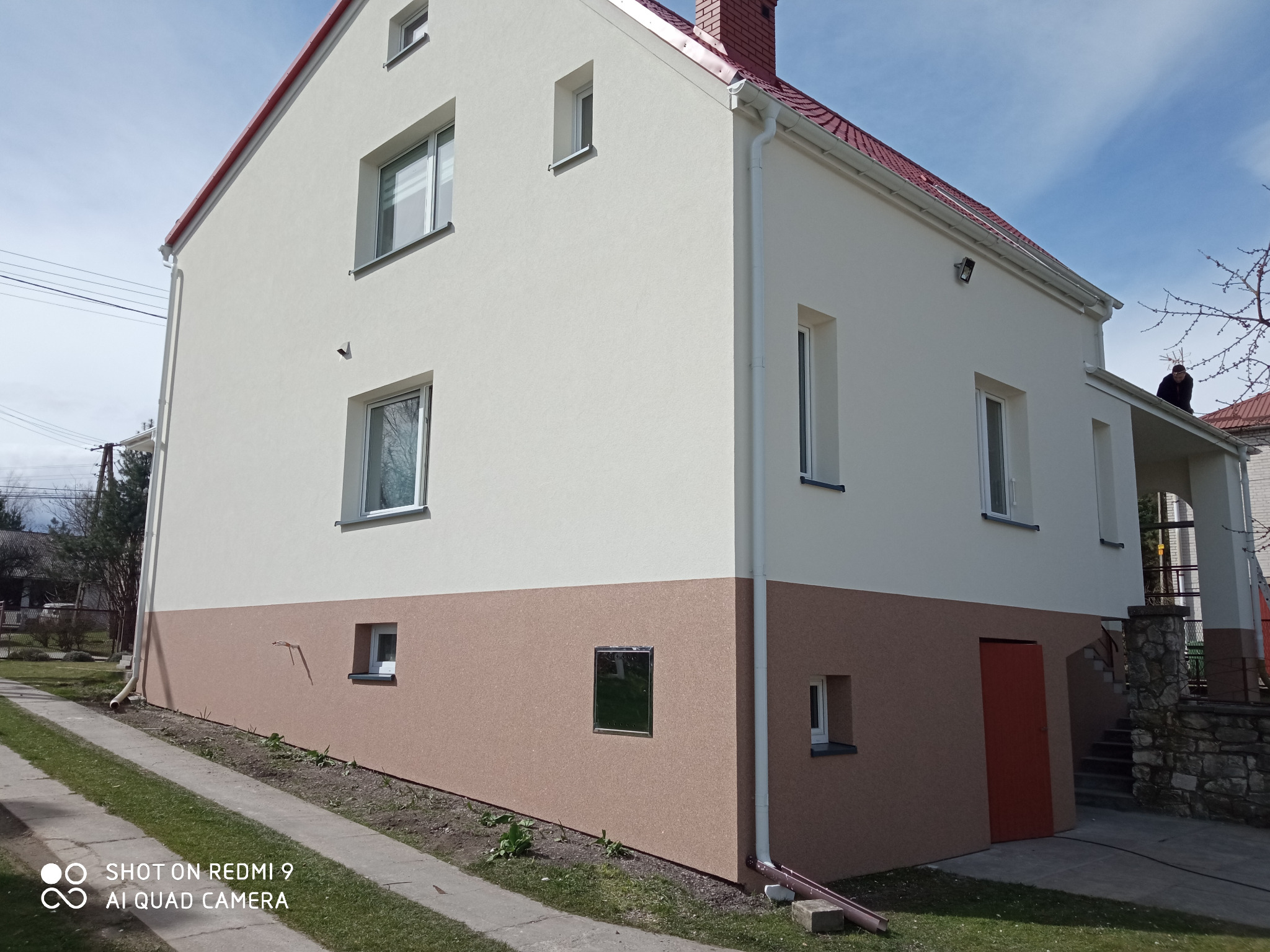 Dwukolorowa elewacja domu jednorodzinnego z czerwoną stolarką dachową, widoczny fragment chodnika i trawnika, ujęcie z poziomu gruntu.