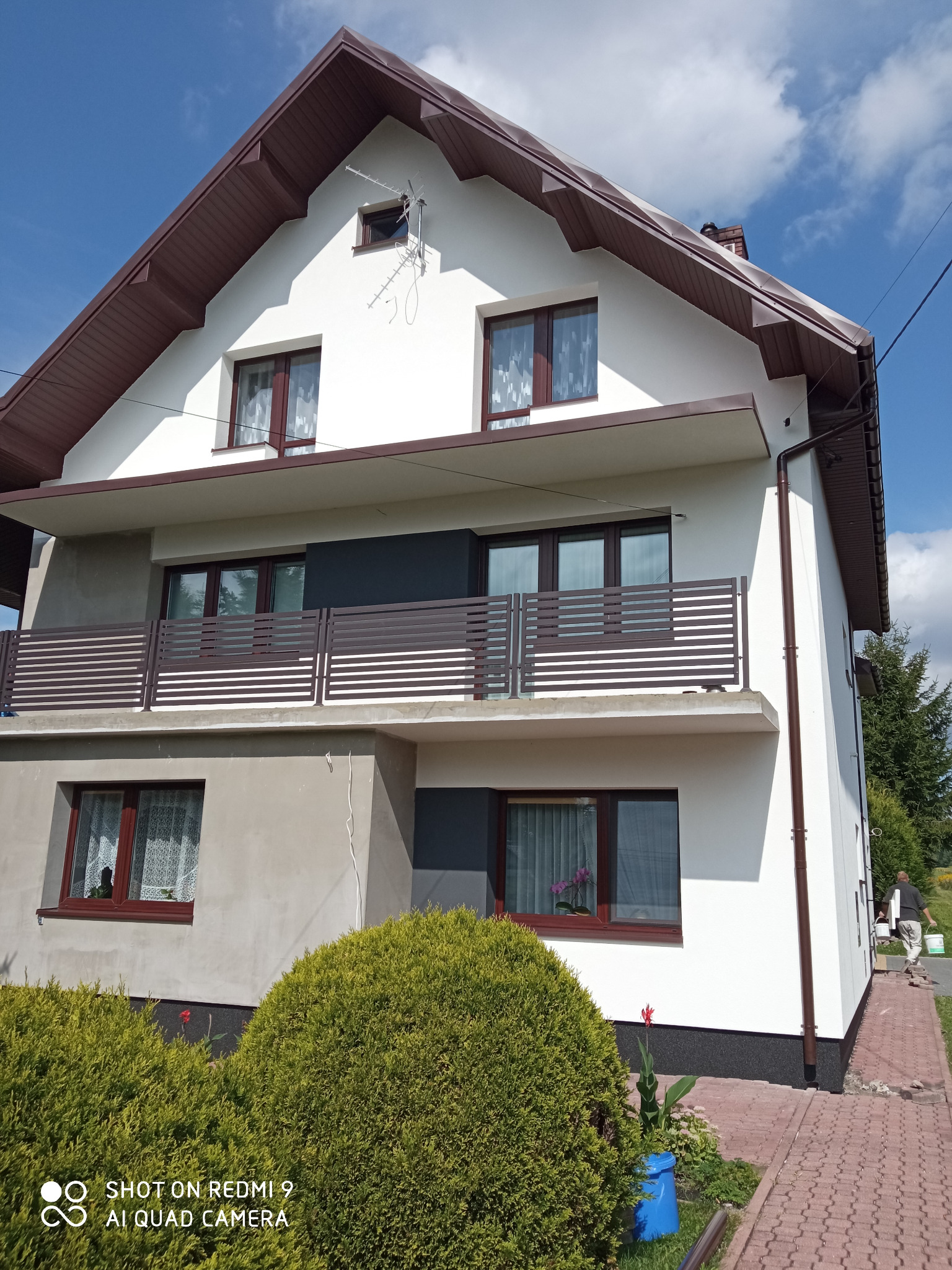 Dwupiętrowy dom jednorodzinny z białą elewacją i brązowym dachem, widoczny po zakończeniu prac, z elementami w kolorze grafitowym, brązowe okna i balustrady, na pierwszym planie zielone krzewy,...