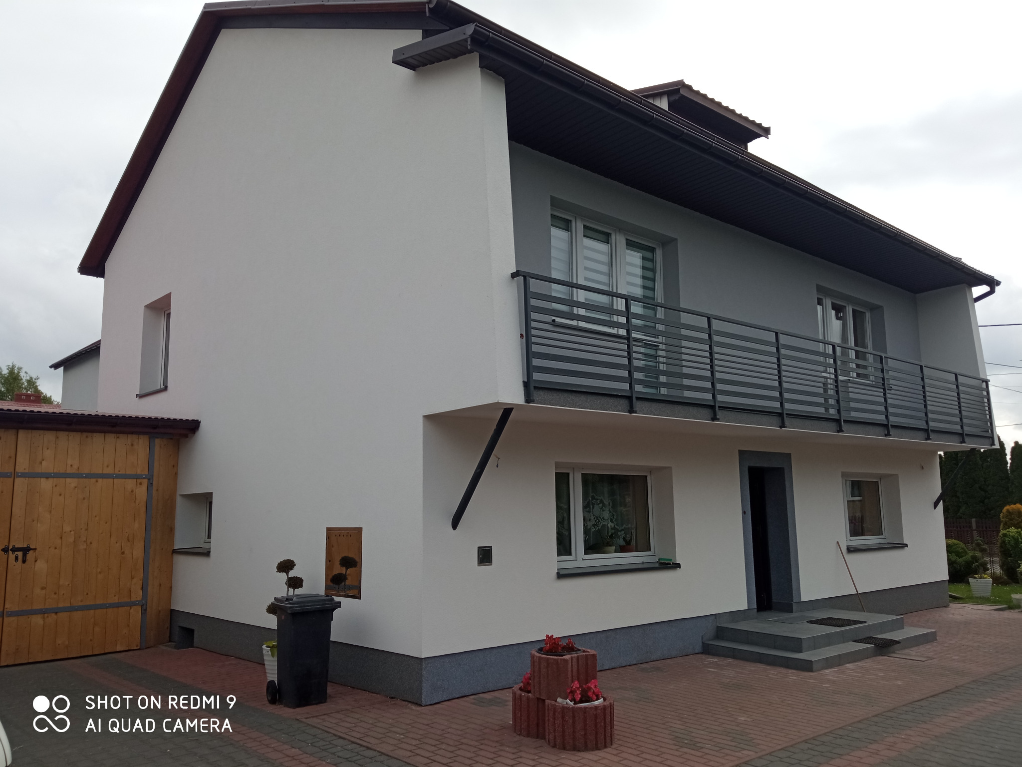 Dwupiętrowy dom z balkonem i szarą elewacją, widok z zewnątrz, z kostką brukową na podjeździe, z drewnianą bramą garażową po lewej stronie i dekoracyjnymi donicami z kwiatami przed wejściem.