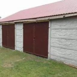 Szary garaż z prefabrykatów betonowych z dwoma brązowymi bramami przesuwnymi, widok z zewnątrz na tle trawy i ogrodzenia.