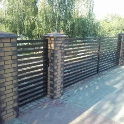 Nowoczesna brama wjazdowa z poziomych, ciemnych profili, osadzona na murowanych słupkach z cegły klinkierowej, zwieńczonych brązowymi daszkami, na tle zieleni.