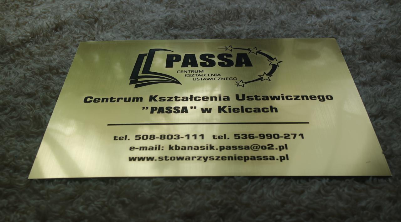 Złota tabliczka informacyjna z czarnym nadrukiem logo i danymi kontaktowymi Centrum Kształcenia Ustawicznego PASSA w Kielcach, leżąca na teksturowanym tle.