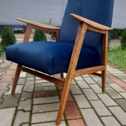 Odnowiony fotel w stylu vintage z drewnianymi podłokietnikami i granatową tapicerką welurową, ustawiony na brukowanym podwórku.
