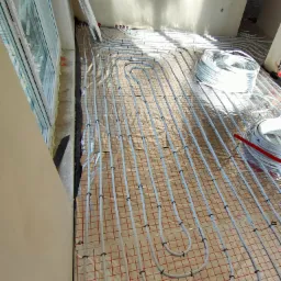 Białe rury systemu rozprowadzenia ciepła ułożone na folii z czerwoną siatką, mocowane czarnymi uchwytami, przygotowane do zalania wylewką.