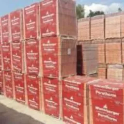 Palety czerwonych bloczków ceramicznych Porotherm, ułożone w stosy na zewnątrz, częściowo owinięte folią. Widoczny znak firmowy producenta.
