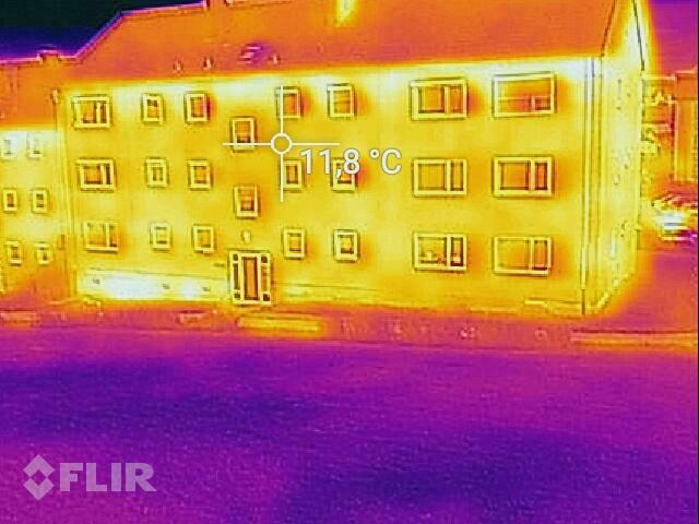 Termowizyjne zdjęcie budynku wielorodzinnego; widoczne różnice temperatur na ścianach i oknach, wskazujące na potencjalne straty ciepła. Na zdjęciu widoczna temperatura 11.8 stopni Celsjusza.