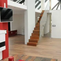 Nowoczesne wnętrze z drewnianymi schodami o minimalistycznym designie, szklana balustrada, jasna podłoga i duże okna dachowe w tle.