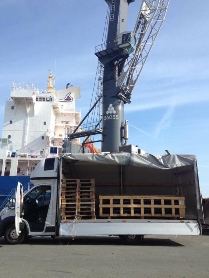 Ciężarówka z otwartą przestrzenią ładunkową załadowana drewnianymi paletami, na tle nabrzeża z dźwigiem Liebherr i częściowo widocznym statkiem towarowym.