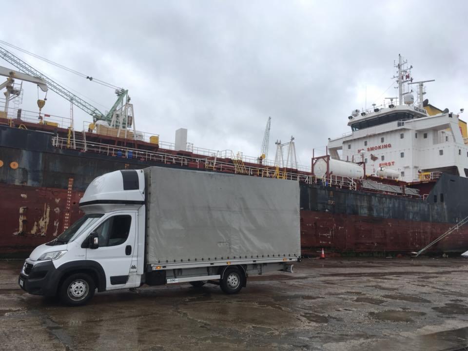 Biały samochód dostawczy na tle zacumowanego statku towarowego z widocznymi napisami ostrzegawczymi, dźwigami portowymi i pochmurnym niebem.