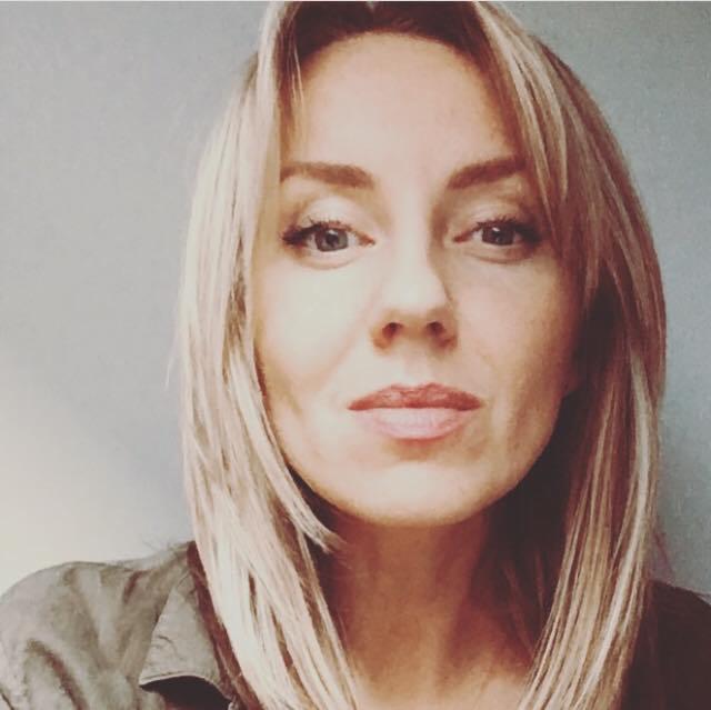 Portret kobiety z blond włosami i zielonej koszuli na tle szarej ściany. Uśmiechająca się twarz, naturalny makijaż, bezpośrednie spojrzenie w obiektyw.