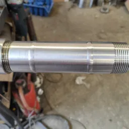 Metalowy wałek z gwintem i widocznym spawem, leżący poziomo. W tle narzędzia i elementy warsztatu.