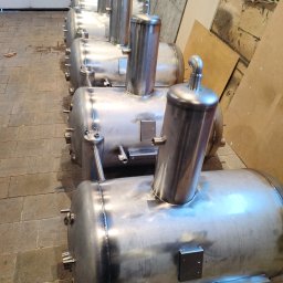 Welder - Usługi Spawalnicze Marcin Kozłowski - Rząd identycznych, spawanych, metalowych zbiorników ciśnieniowych z przyłączami, ustawionych na posadzce z płytek. Widoczne spawy i elementy montażowe.