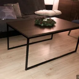 Nowoczesny stolik kawowy z ciemnego drewna z czarnymi, metalowymi nogami, udekorowany świątecznym stroikiem na tle sofy z poduszkami