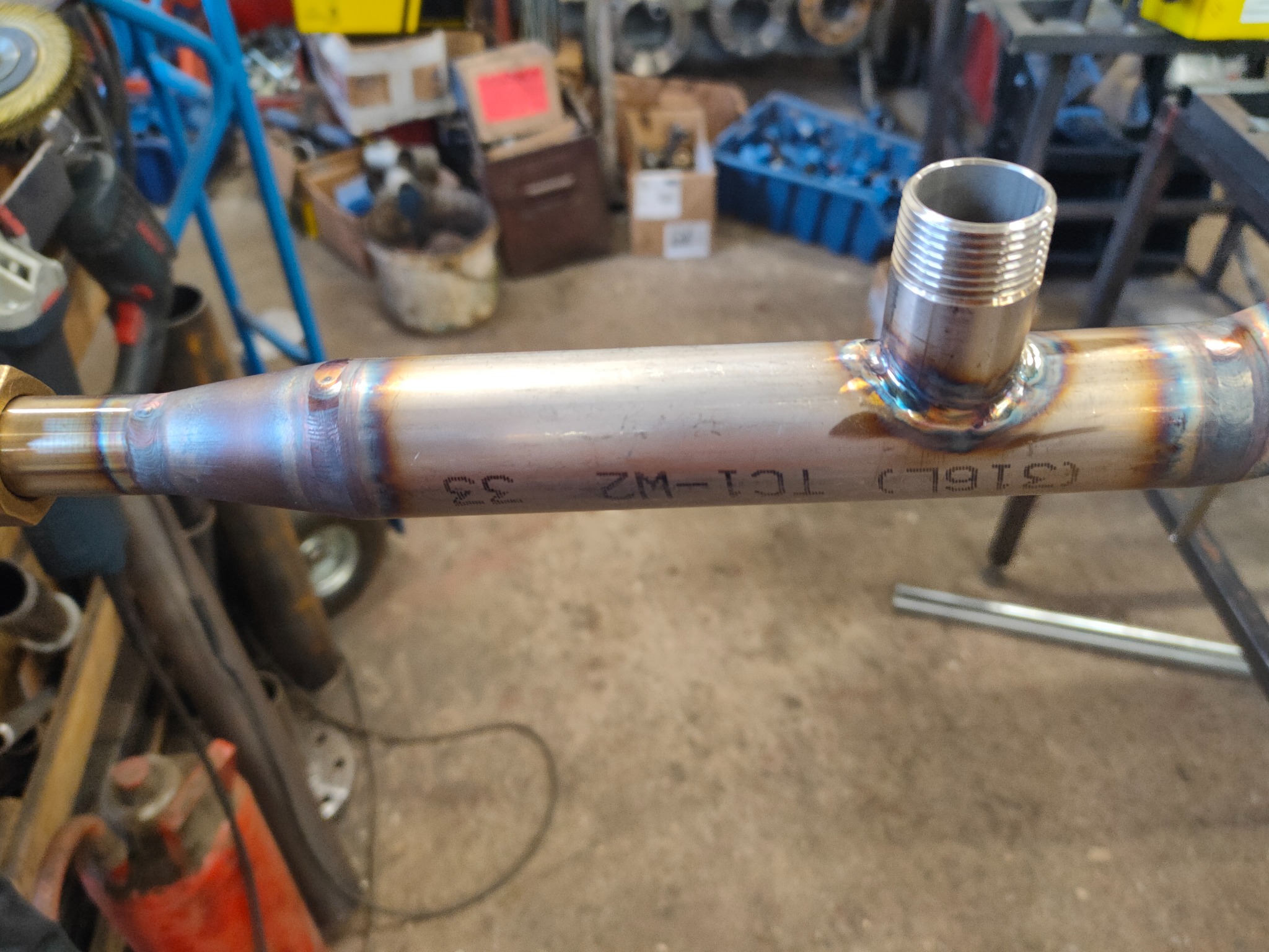 Metalowa rura z przyspawanym gwintem, widoczne ślady po spawaniu i oznaczenia (316L) TC1-WZ 33, w tle warsztat z narzędziami.