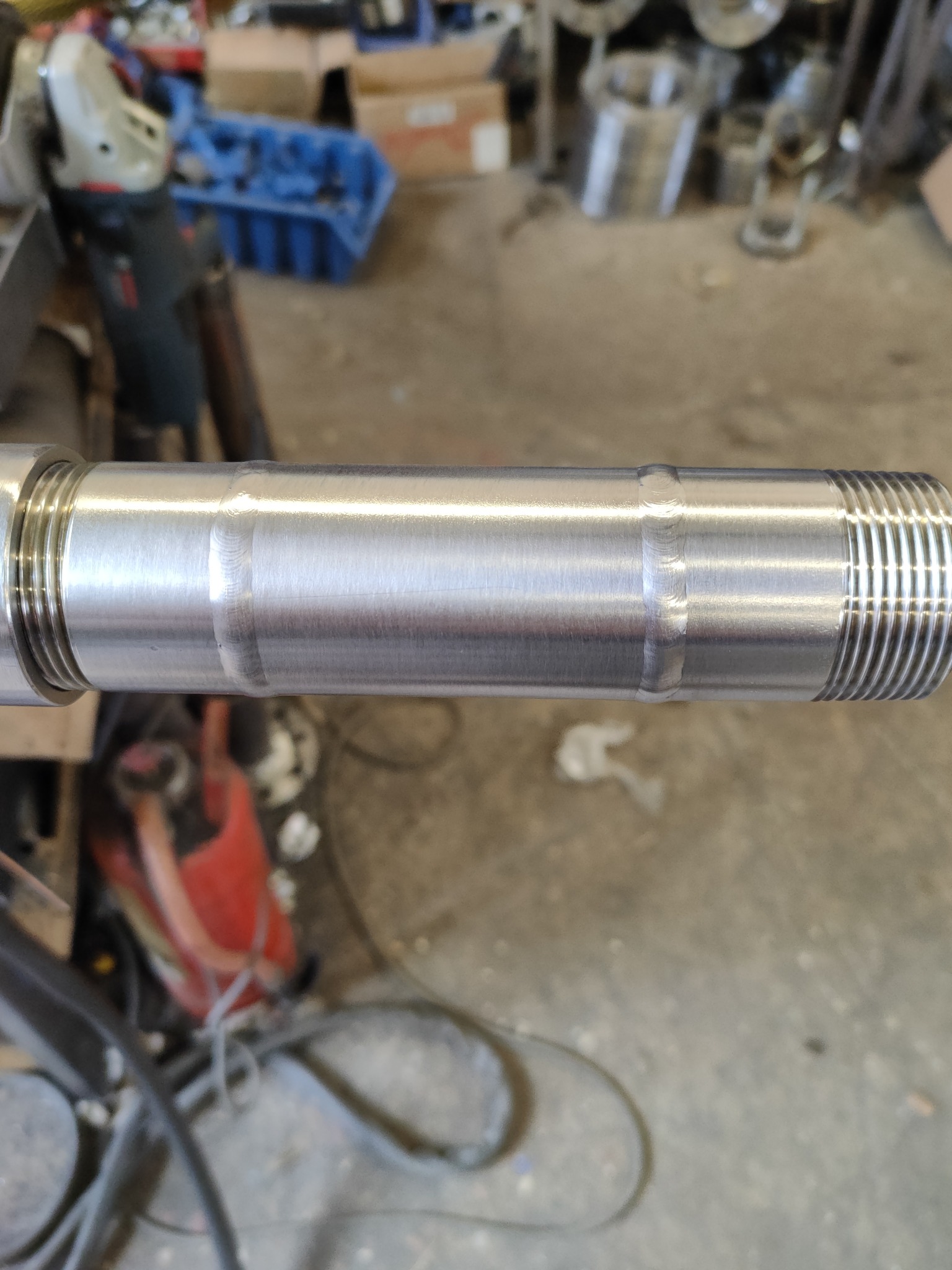 Metalowy wałek z gwintem i widocznym spawem, leżący poziomo. W tle narzędzia i elementy warsztatu.