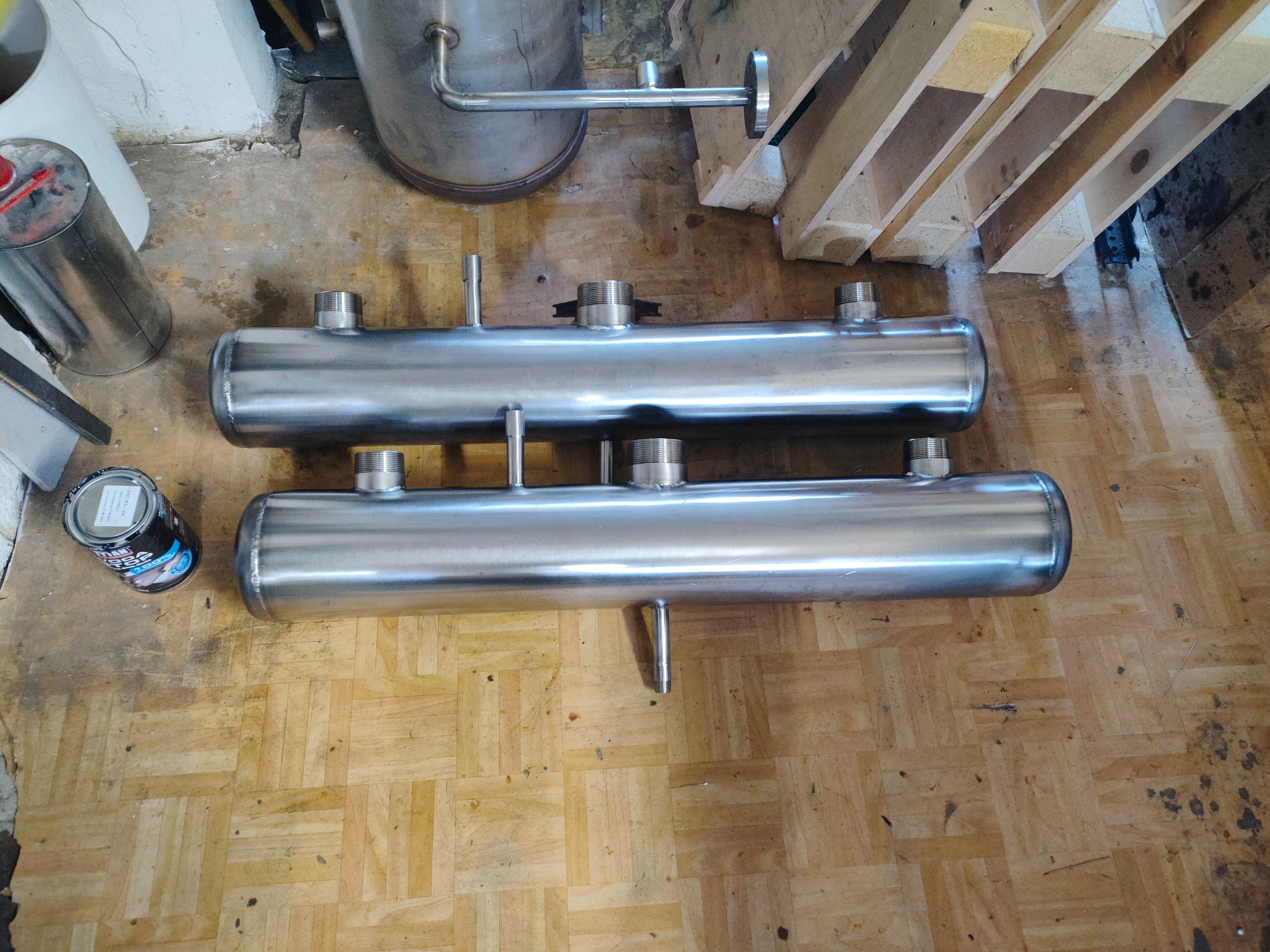 Dwa identyczne, spawane zbiorniki ze stali nierdzewnej z gwintowanymi króćcami, leżące na parkiecie. Obok puszka farby i elementy konstrukcji drewnianej.