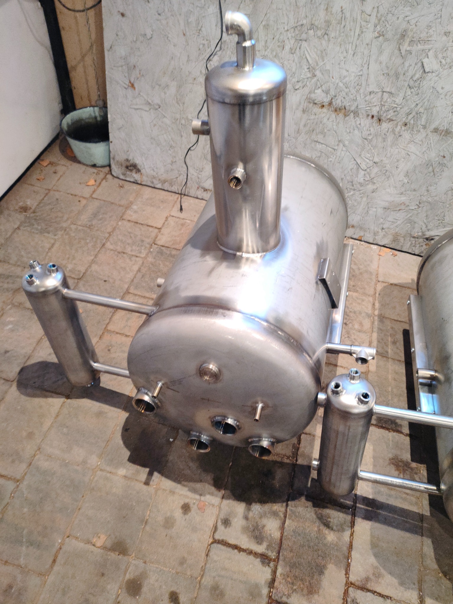 Zbiornik ciśnieniowy ze stali nierdzewnej z widocznymi spawami i elementami rurowymi na tle płytek chodnikowych i ściany. Precyzyjne wykonanie.