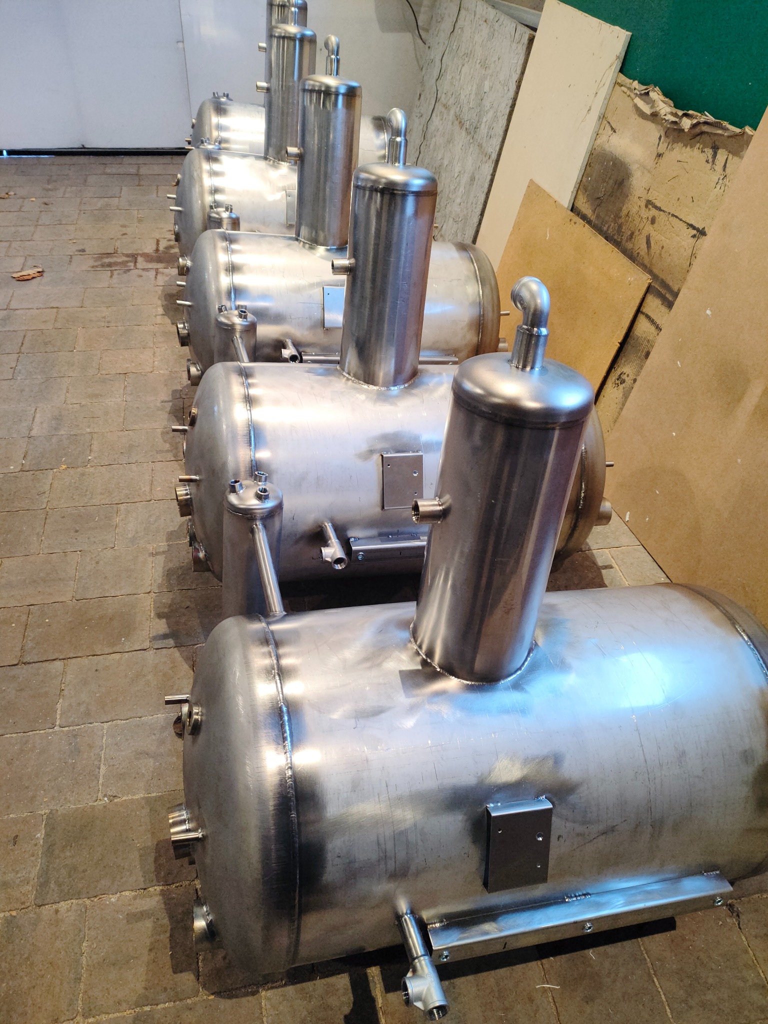 Rząd identycznych, spawanych, metalowych zbiorników ciśnieniowych z przyłączami, ustawionych na posadzce z płytek. Widoczne spawy i elementy montażowe.
