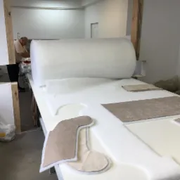 Przygotowywanie elementów z materiału i pianki na dużym stole w szwalni, widoczny wałek z pianką, wykroje i pracownik przy pracy.