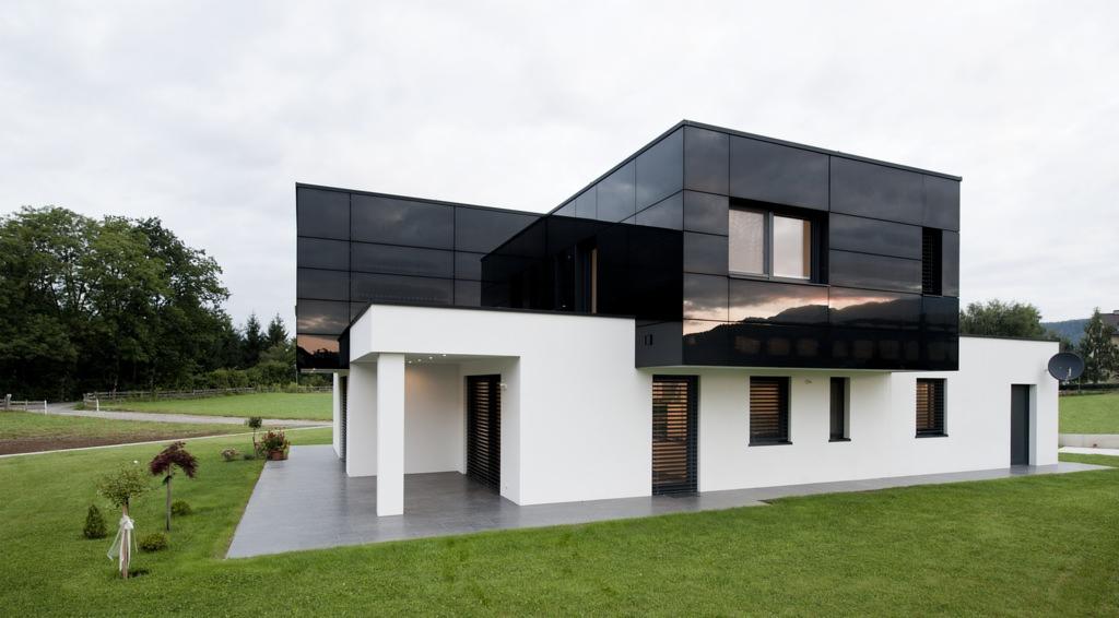 Nowoczesny dom z czarną, lustrzaną elewacją i białymi ścianami, otoczony zielonym trawnikiem i młodymi drzewkami, z widocznym odbiciem gór w powierzchni elewacji.