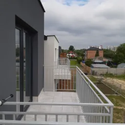 Nowoczesny balkon z szarą elewacją, jasnymi płytkami i metalową balustradą, widok na okoliczne budynki i pochmurne niebo.