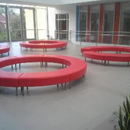 Hol z szarymi płytkami podłogowymi i okrągłymi, czerwonymi sofami modułowymi na metalowych n&oacute;żkach, widok z perspektywy rośliny doniczkowej w rogu.