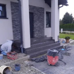 Pracownik w trakcie układania szarych płytek elewacyjnych na schodach wejściowych do domu, widoczny kubeł z narzędziami, odkurzacz przemysłowy i paleta z płytkami.