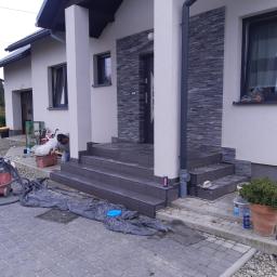 RZETELNA AKCJA PAWEL RÓZGA - Układanie płytek na schodach wejściowych do domu jednorodzinnego, widoczny pracownik w trakcie pracy, narzędzia, odkurzacz przemysłowy, na podjeździe rozłożona folia zabezpieczająca.