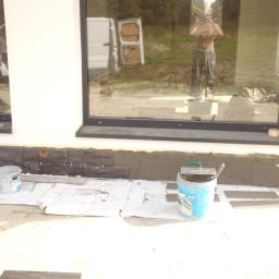 RZETELNA AKCJA PAWEL RÓZGA - Montaż ciemnych płytek elewacyjnych przy oknie, widoczne narzędzia i zaprawa, w tle odbicie osoby robiącej zdjęcie i samochód dostawczy.