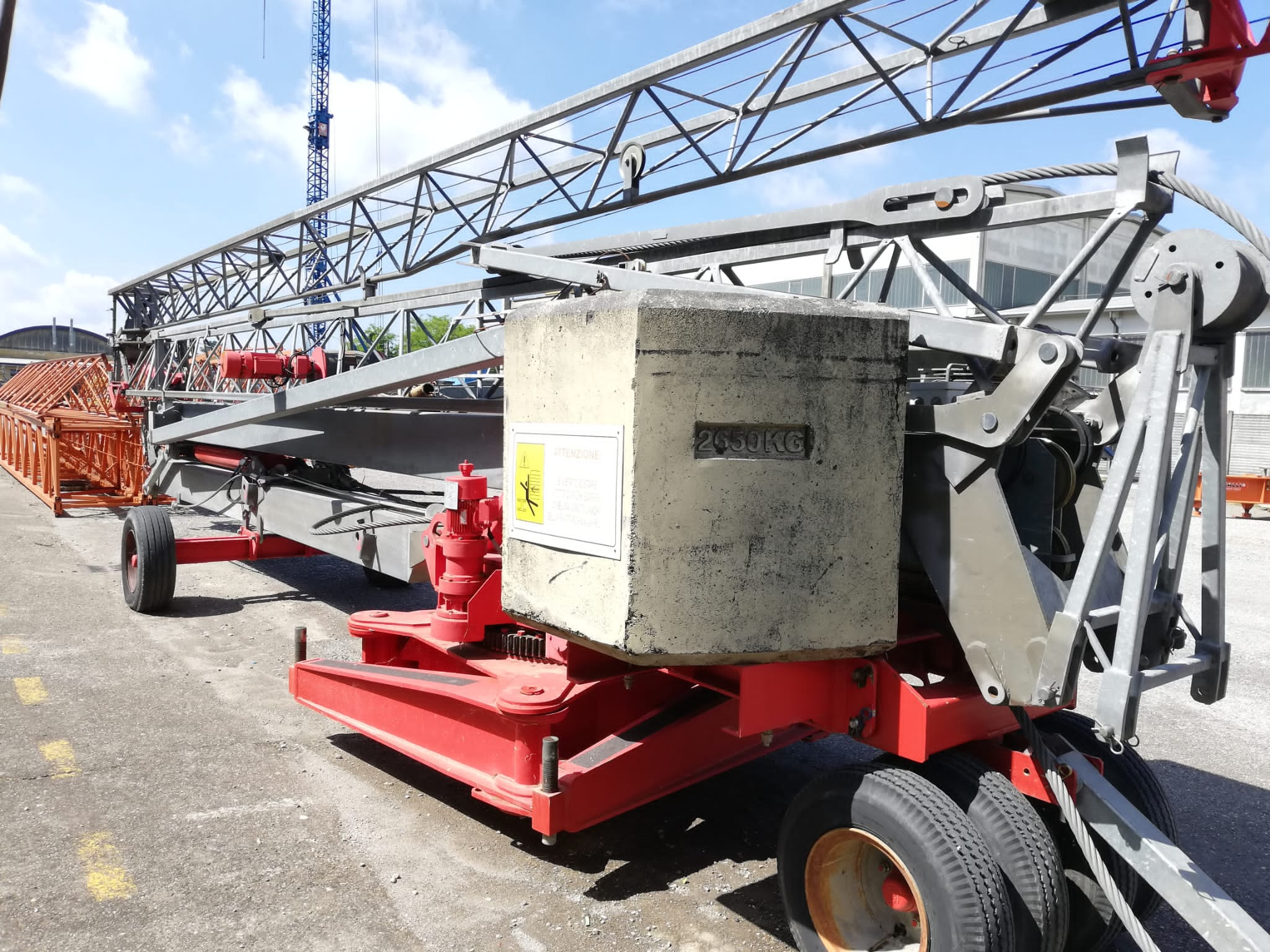 Przenośny żuraw wieżowy z betonowym balastem 2650 kg i wysuwanym ramieniem kratownicowym, widoczny w tle inny żuraw wieżowy.