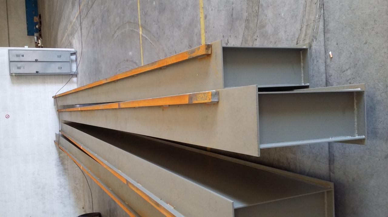 Stalowe belki H ułożone warstwami w hali produkcyjnej, widoczne ślady rdzy na krawędziach belek, szara betonowa podłoga i ściany w tle.