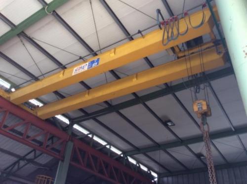 Żółty suwnicowy dźwig pomostowy zamontowany pod dachem hali, widoczne łańcuchy i wciągarka