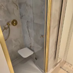 Nowoczesna kabina prysznicowa z marmurowymi płytkami, złotymi detalami i siedziskiem, widoczne zabezpieczenie podłogi kartonem.